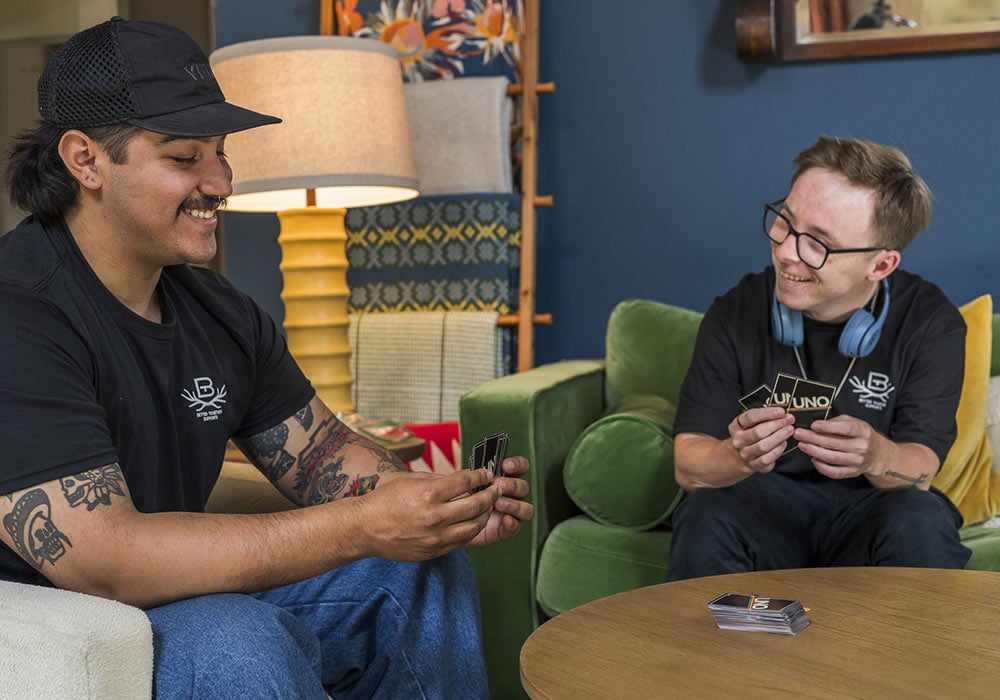 At right, a special needs young man in glasses with headphones around his neck, plays cards with a direct support professional staffer wearing a baseball cap