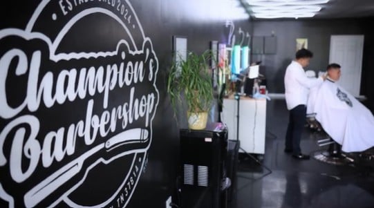 The interior of a barbershop shows a barber cutting a client’s hair beside a wall featuring a large ‘Champion’s Barbershop’ logo, with mirrors, plants, and grooming stations in the background.