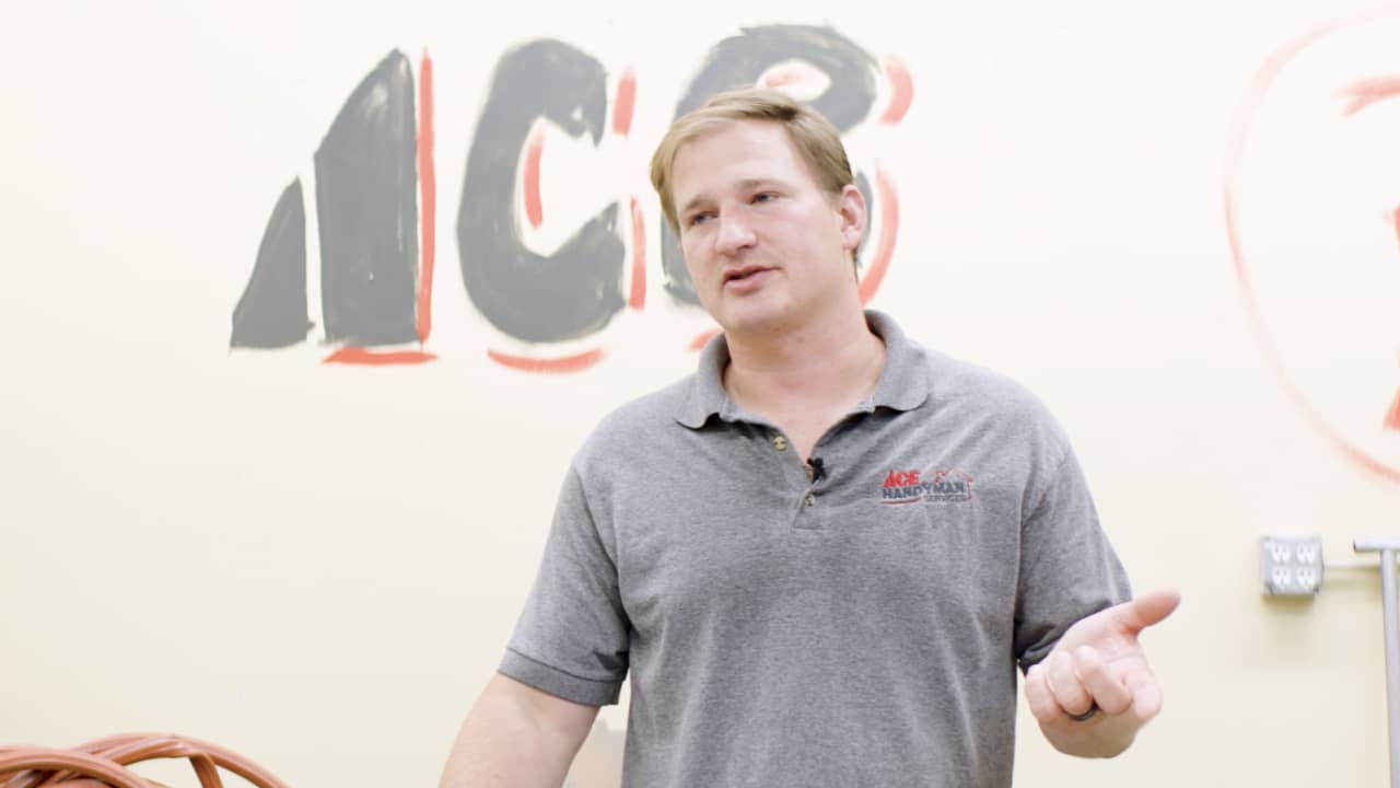 man in grey shirt talks in front of wall with ACE Handyman Services logo